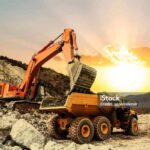 Excavator loading dumper truck on mining site at sunset.